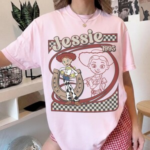 May include: A light pink t-shirt featuring a graphic of the character Jessie from Toy Story. The design includes the name "Jessie" above the image, the year "1995", and a horseshoe. The shirt has a classic crew neck.