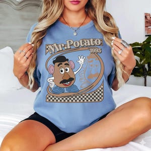 May include: A light blue t-shirt featuring a graphic of Mr. Potato Head with the text "Mr. Potato 1995". The design includes a cartoon image of Mr. Potato Head waving, with a brown and black checkered border at the bottom.