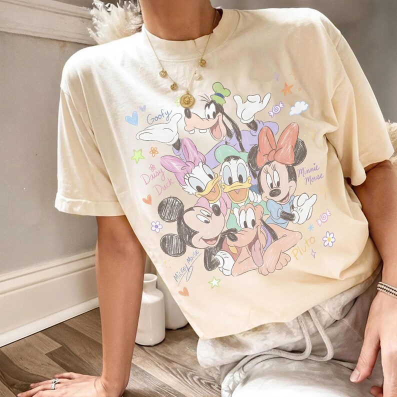 May include: Cream-colored t-shirt featuring a colorful graphic of Disney characters including Mickey Mouse, Minnie Mouse, Donald Duck, Daisy Duck, Goofy, and Pluto. The design includes the characters' names and other decorative elements.