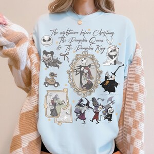 May include: Pale blue sweatshirt with a design from "The Nightmare Before Christmas." The graphic includes Jack Skellington, Sally, and other characters in a collage. The text reads "The Nightmare Before Christmas & The Pumpkin Queen & The Pumpkin King."
