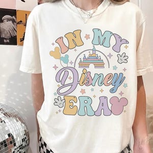 Op de afbeelding: Cr&egrave;mekleurig T-shirt met een kleurrijke afbeelding met de tekst "IN MY Disney ERA". Het ontwerp bevat een kasteel, Mickey Mouse-handen, ballonnen en sterren in pastelkleuren. Een casual, comfortabel T-shirt.
