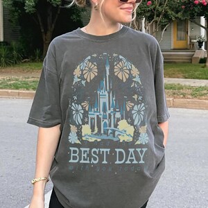 May include: Dark gray t-shirt featuring a castle design with floral accents in blue, tan, and white. The text "I had the BEST DAY with you today" is printed below the castle image. The shirt has short sleeves.
