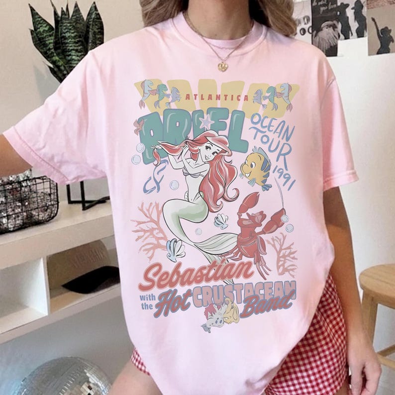 May include: Light pink t-shirt featuring a graphic of Ariel from The Little Mermaid, with the text "Ariel Ocean Tour 1991." The design includes Ariel, Flounder, Sebastian, and coral. The shirt is a crew neck.