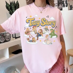 May include: A light pink short-sleeve t-shirt featuring a graphic print with the text Tay Story in yellow letters. The design includes multiple illustrated figures dressed in various costumes sitting on clouds, surrounded by yellow stars and music notes.