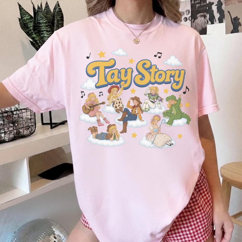 May include: A light pink short-sleeve t-shirt featuring a graphic print with the text Tay Story in yellow letters. The design includes multiple illustrated figures dressed in various costumes sitting on clouds, surrounded by yellow stars and music notes.