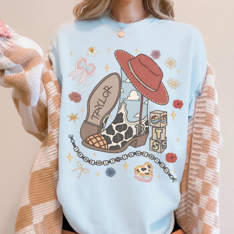 May include: A light blue crewneck sweatshirt featuring a graphic print of a cow-print cowboy boot, a red hat, blocks with the letters T, S, and a heart, and a beaded string with the text I KNEW IT Y I KNEW YOU. The boot has the name TAYLOR.