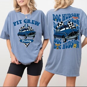 May include: A pale blue t-shirt with a graphic of a blue car and checkered flags. The front reads "PIT CREW Daddy", and the back says "DOC HUDSON Cars". The shirt has a loose fit and short sleeves, suitable for casual wear.