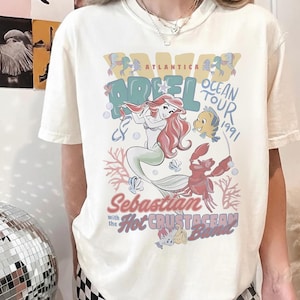 May include: Cream-colored t-shirt featuring a colorful graphic of Ariel from The Little Mermaid. The design includes Ariel, Flounder, Sebastian, and the text "Ariel Ocean Tour 1991" and "Hot Crustacean Band".