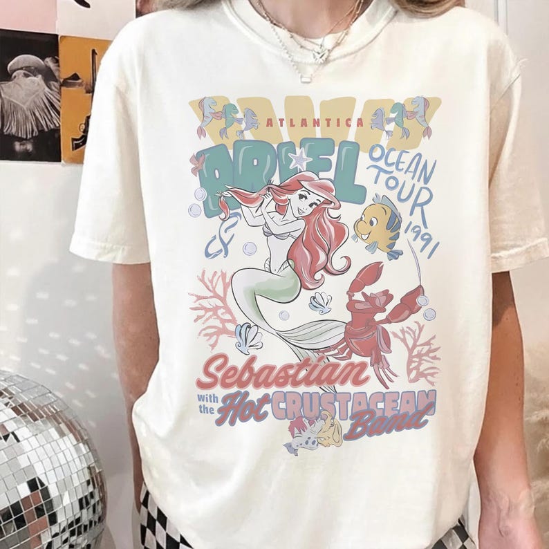 May include: Cream-colored t-shirt featuring a colorful graphic of Ariel from The Little Mermaid. The design includes Ariel, Flounder, Sebastian, and the text "Ariel Ocean Tour 1991" and "Hot Crustacean Band".