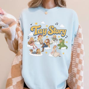 May include: A light blue crewneck sweatshirt featuring the text Tay Story in yellow with a blue outline. The graphic includes several illustrated figures dressed in various costumes sitting on white clouds, accented by yellow stars and musical notes.