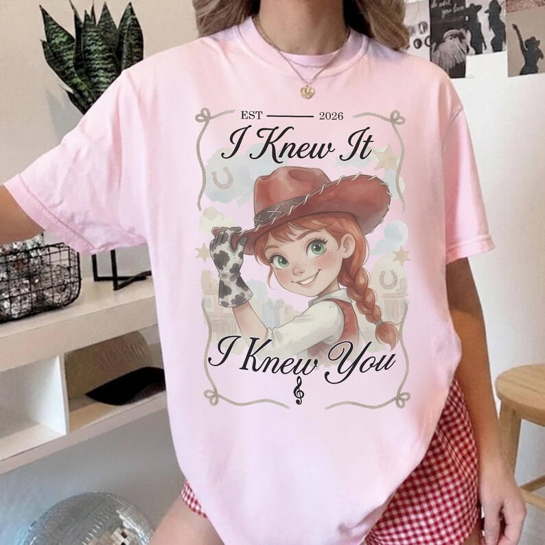 May include: A light pink t-shirt featuring a graphic of a cartoon cowgirl with red hair in braids and a brown hat. The text reads EST 2026, I Knew It, and I Knew You, framed by a decorative border with stars and horseshoes on a light background.