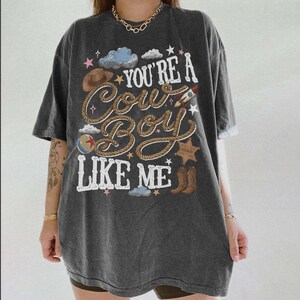 Puede incluir: Una camiseta de manga corta gris carb&oacute;n con un estampado gr&aacute;fico que incluye el texto "YOU'RE A COWBOY LIKE ME" en letras blancas. El dise&ntilde;o presenta ilustraciones de un sombrero de vaquero marr&oacute;n, una cuerda, un cohete, una pelota, una estrella de sheriff y botas.