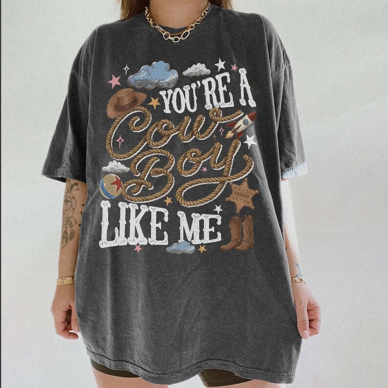 Puede incluir: Una camiseta de manga corta gris carb&oacute;n con un estampado gr&aacute;fico que incluye el texto "YOU'RE A COWBOY LIKE ME" en letras blancas. El dise&ntilde;o presenta ilustraciones de un sombrero de vaquero marr&oacute;n, una cuerda, un cohete, una pelota, una estrella de sheriff y botas.