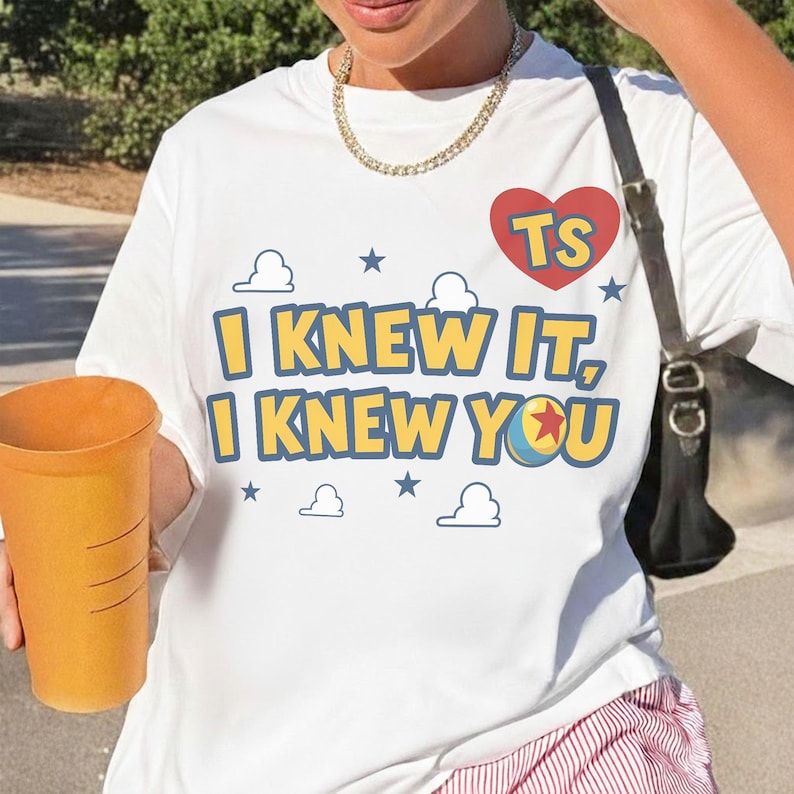 May include: A white short-sleeved t-shirt featuring the text I KNEW IT, I KNEW YOU in yellow with blue outlines. The design includes a red heart with the letters TS, blue stars, white cloud outlines, and a colorful ball with a red star.
