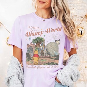 Puede incluir: Una camiseta de color lila claro con un dise&ntilde;o de Disney World. El dise&ntilde;o incluye el texto "Florida Disney World Est. 1971" y una ilustraci&oacute;n de Winnie the Pooh, el Hollywood Tower Hotel y Spaceship Earth. La camiseta tambi&eacute;n tiene el texto "The Most Magical Place On Earth."