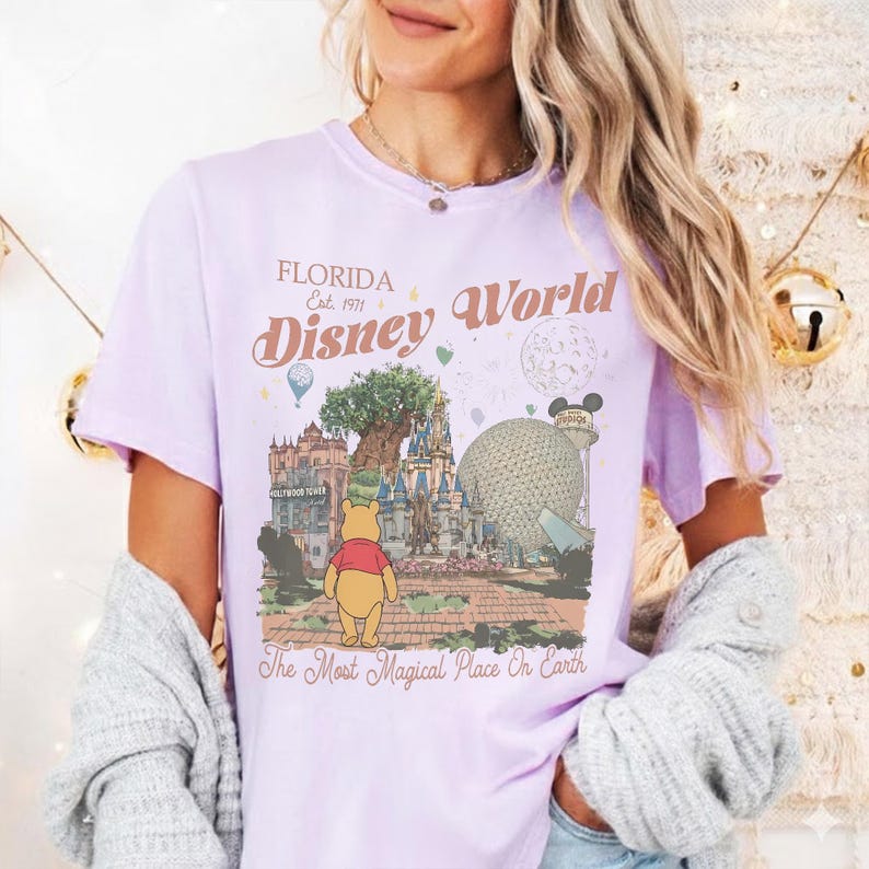 Puede incluir: Una camiseta de color lila claro con un dise&ntilde;o de Disney World. El dise&ntilde;o incluye el texto "Florida Disney World Est. 1971" y una ilustraci&oacute;n de Winnie the Pooh, el Hollywood Tower Hotel y Spaceship Earth. La camiseta tambi&eacute;n tiene el texto "The Most Magical Place On Earth."
