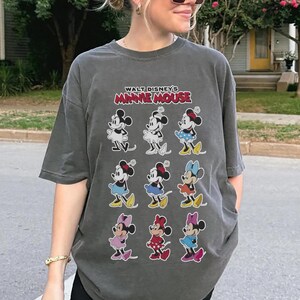 K&ouml;nnte beinhalten: Dunkelgraues T-Shirt mit mehreren Minnie Mouse-Illustrationen in verschiedenen Outfits. Der Text "WALT DISNEY'S MINNIE MOUSE" ist in Rot und Wei&szlig; &uuml;ber den Bildern aufgedruckt. Das Shirt hat einen Vintage-Look.
