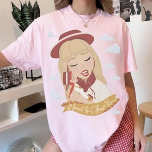 May include: A light pink short-sleeve t-shirt featuring a graphic illustration of a blonde character wearing a red hat, holding a smaller doll, surrounded by white clouds. Below the illustration is a banner with the text I Knew It, I Knew You.