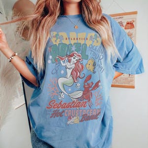 May include: Light blue t-shirt featuring a graphic of Ariel from The Little Mermaid. The design includes Ariel, Flounder, Sebastian, and the text "Atlantica Ocean Tour 1991". The shirt has short sleeves and a relaxed fit.
