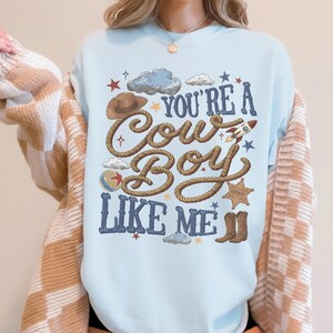 Puede incluir: Una sudadera de cuello redondo en color azul claro con un estampado gr&aacute;fico que incluye el texto "YOU'RE A COWBOY LIKE ME" en letras azul oscuro. El dise&ntilde;o presenta ilustraciones de un sombrero de vaquero marr&oacute;n, una cuerda, un cohete, una pelota, una estrella de sheriff y botas.