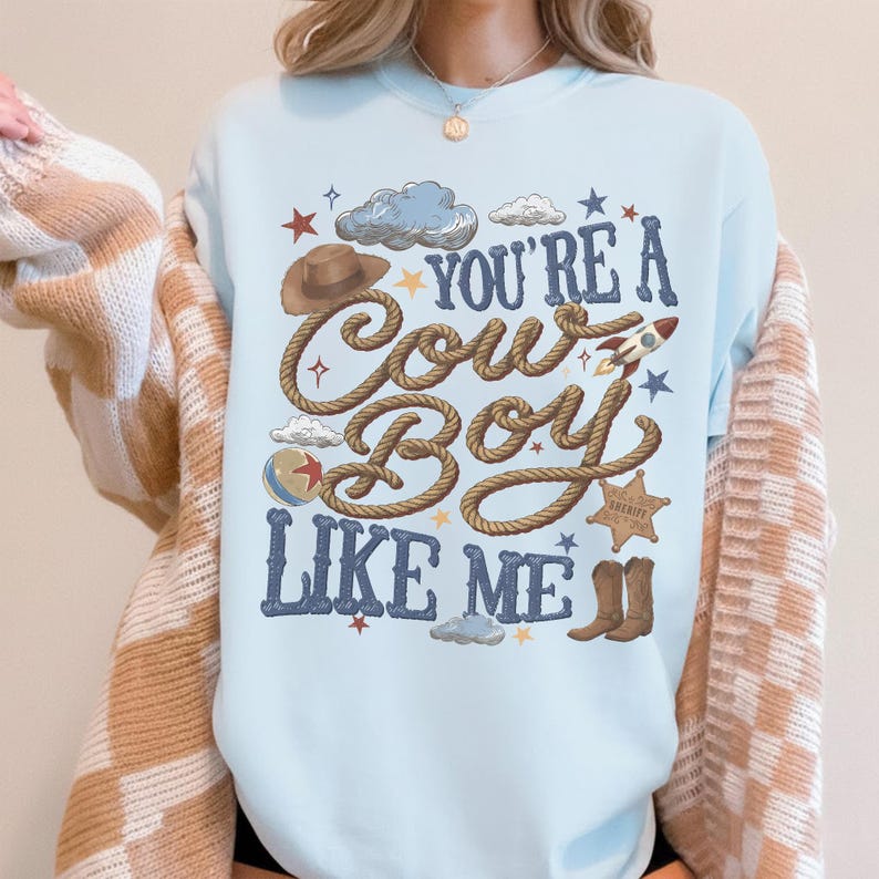 Puede incluir: Una sudadera de cuello redondo en color azul claro con un estampado gr&aacute;fico que incluye el texto "YOU'RE A COWBOY LIKE ME" en letras azul oscuro. El dise&ntilde;o presenta ilustraciones de un sombrero de vaquero marr&oacute;n, una cuerda, un cohete, una pelota, una estrella de sheriff y botas.