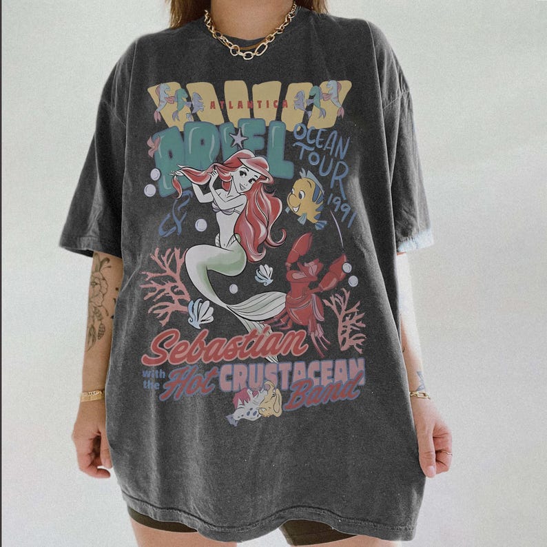 May include: Dark gray t-shirt featuring a graphic of Ariel from The Little Mermaid, with the text "Atlantica Hotel Ocean Tour 1991" and "Sebastian with the Hot Crustacean Band". The design includes coral, fish, and other sea elements.