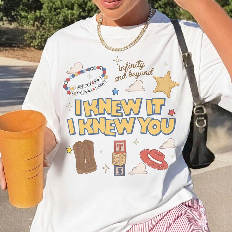 May include: A white short-sleeve t-shirt featuring a graphic print with the text "I KNEW IT I KNEW YOU", "infinity and beyond", and "YOU'VE GOT A FRIEND IN ME". The design includes illustrations of cowboy boots, a hat, blocks, and a beaded bracelet.