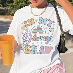 Op de afbeelding: Wit T-shirt met de tekst "IN MY Disney ERA" in pastelkleuren. Het ontwerp bevat een kasteel, Mickey Mouse-handen en sterren. Het shirt is een casual stijl.
