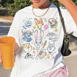 Op de afbeelding: Wit T-shirt met een kleurrijk cartoonontwerp met personages uit een film. Het ontwerp bevat Judy Hopps, Nick Wilde, Flash en anderen. De namen van de personages staan op het shirt.