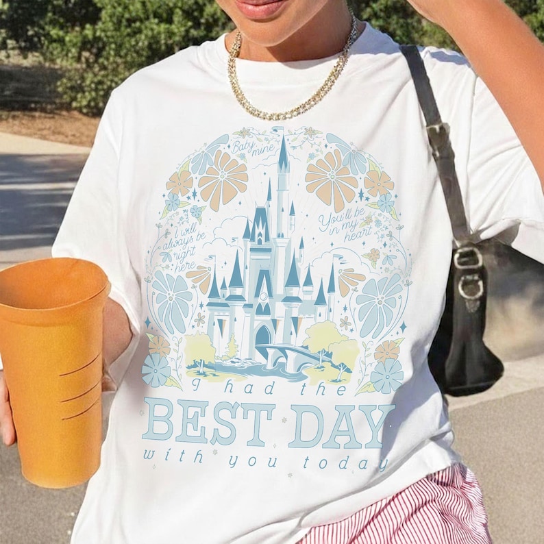 May include: White t-shirt featuring a whimsical castle design in shades of blue, yellow, and beige, surrounded by floral elements and the text "I had the BEST DAY with you today." The shirt has a crew neck and short sleeves.