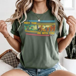 May include: Olive green t-shirt featuring the text "MAY THE FOURTH BE WITH YOU" in a retro style. The design incorporates blue, yellow, and red light saber graphics. The sleeves of the shirt are rolled up.
