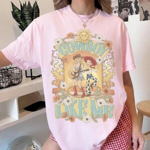 May include: A light pink t-shirt featuring a graphic print of two cartoon characters, one playing a guitar, surrounded by flowers and a sun. The text "COWBOY LIKE ME" is printed in large, light blue letters above and below the central illustration.