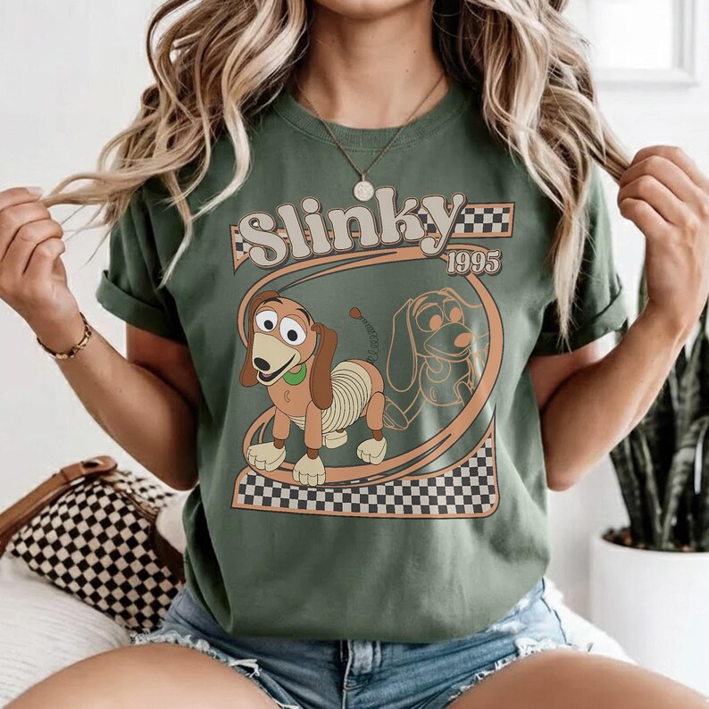 May include: Olive green t-shirt featuring a graphic of Slinky Dog from Toy Story. The design includes the word "Slinky" above the dog, with "1995" below. The graphic is framed by a checkered pattern.