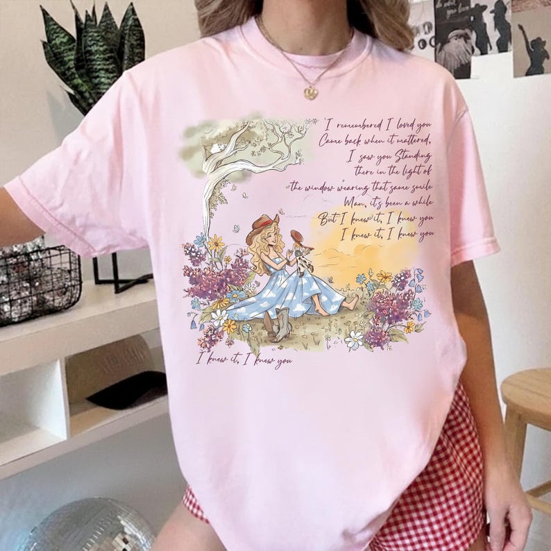 May include: A light pink short-sleeve t-shirt featuring a graphic of a person in a blue dress and cowboy boots sitting in a field of flowers under a tree. The text reads: I remembered I loved you, Came back when it mattered, I saw you standing there in the light of the window wearing that same smile, Man, it's been a while, But I knew it, I knew you, I knew it, I knew you.