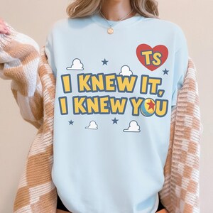 May include: A light blue crewneck sweatshirt featuring a red heart graphic with the letters TS, and the yellow text I KNEW IT, I KNEW YOU. The design includes small white clouds and blue stars, with a colorful ball replacing the letter O.