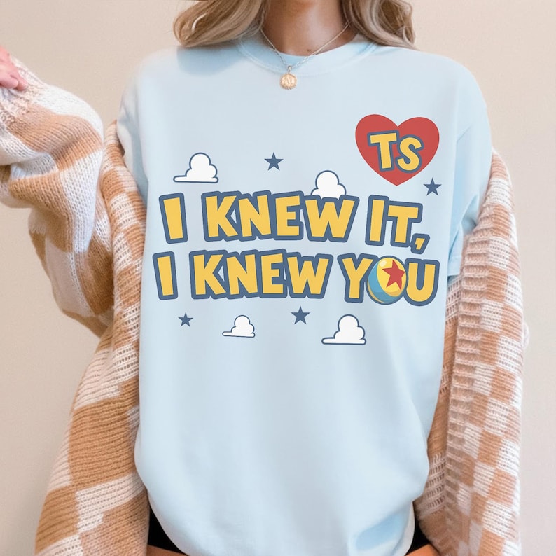 May include: A light blue crewneck sweatshirt featuring a red heart graphic with the letters TS, and the yellow text I KNEW IT, I KNEW YOU. The design includes small white clouds and blue stars, with a colorful ball replacing the letter O.