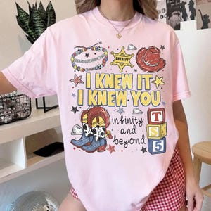 May include: A light pink t-shirt featuring a graphic print with a sheriff badge, a cowboy hat, cow-print boots, and colorful blocks. The text reads I KNEW IT I KNEW YOU, infinity and beyond, and YOU'VE GOT A FRIEND IN ME, with various stars.