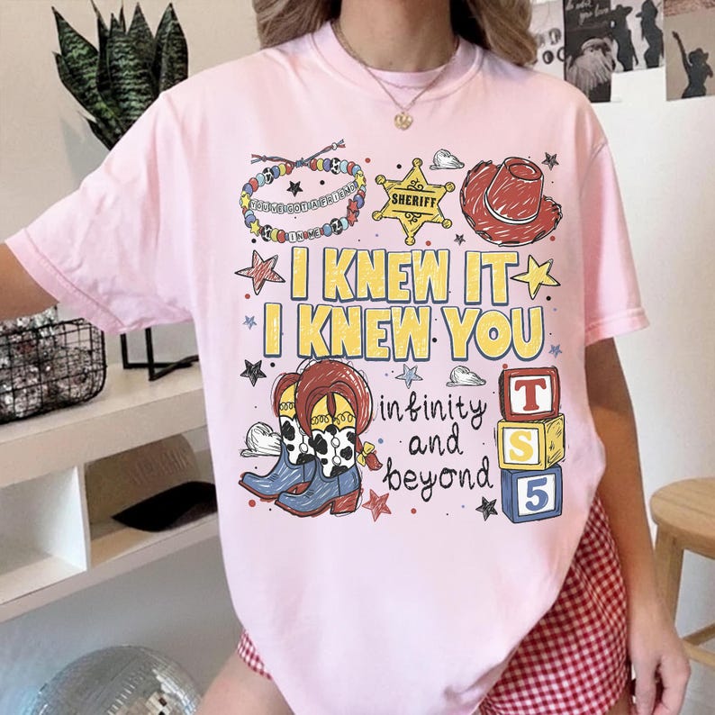 May include: A light pink t-shirt featuring a graphic print with a sheriff badge, a cowboy hat, cow-print boots, and colorful blocks. The text reads I KNEW IT I KNEW YOU, infinity and beyond, and YOU'VE GOT A FRIEND IN ME, with various stars.