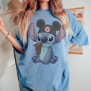 May include: Light blue t-shirt featuring a cartoon Stitch character wearing a Mickey Mouse hat and holding a chocolate ice cream bar. The graphic is centered on the shirt. The shirt has short sleeves and a relaxed fit.