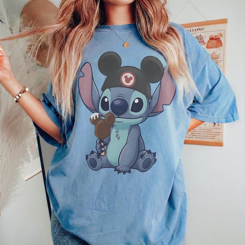May include: Light blue t-shirt featuring a cartoon Stitch character wearing a Mickey Mouse hat and holding a chocolate ice cream bar. The graphic is centered on the shirt. The shirt has short sleeves and a relaxed fit.