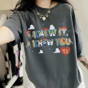 May include: A charcoal gray short-sleeve t-shirt featuring the text I KNEW IT, I KNEW YOU in a colorful, patterned font inspired by Toy Story characters. The design includes small blue stars, white clouds, and a red heart with the letters TS inside.