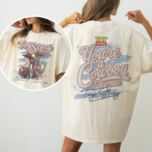 Puede incluir: Una camiseta color crema con un estampado gr&aacute;fico en la espalda con el texto Toy Story, You're a Cowboy like me y Tay Story sobre un dise&ntilde;o de pincelada azul. El gr&aacute;fico frontal muestra dos figuras con atuendo de vaquero y una guitarra.