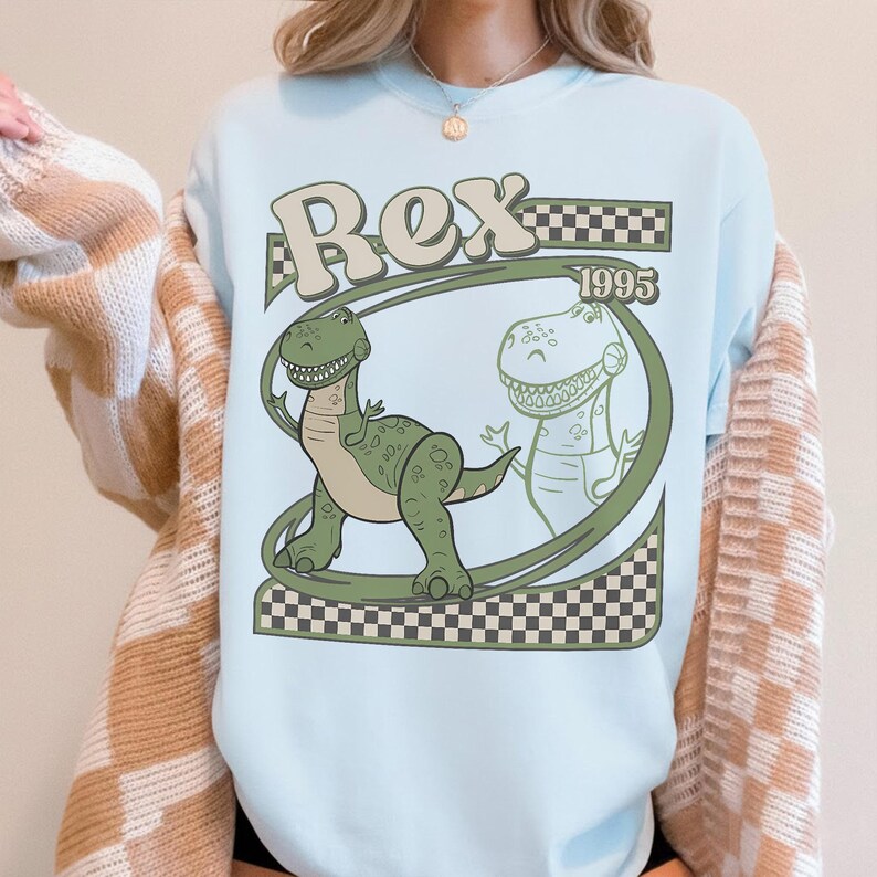 May include: Light blue sweatshirt featuring a cartoon dinosaur named Rex. The design includes the name "Rex" in a retro font, the year "1995", and a checkered pattern. The dinosaur is green with a light green outline.