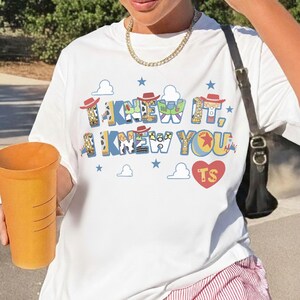 May include: A white short-sleeved t-shirt featuring a graphic print with the text "I KNEW IT, I KNEW YOU" in blue letters filled with character patterns, small red cowboy hats, blue stars, white clouds, and a red heart containing the letters "TS".