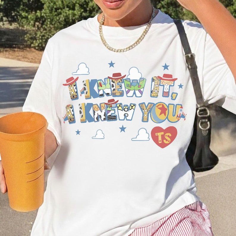 May include: A white short-sleeved t-shirt featuring a graphic print with the text "I KNEW IT, I KNEW YOU" in blue letters filled with character patterns, small red cowboy hats, blue stars, white clouds, and a red heart containing the letters "TS".