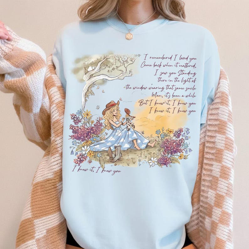 May include: A light blue crewneck sweatshirt featuring a graphic illustration of a person with long blonde hair sitting in a field of flowers under a tree. The design includes the text: I remembered I loved you, Come back when it mattered, I saw you standing there in the light of the window wearing that same smile, Man, it's been a while, But I knew it, I knew, you I knew it, I knew you, I knew it, I knew you.