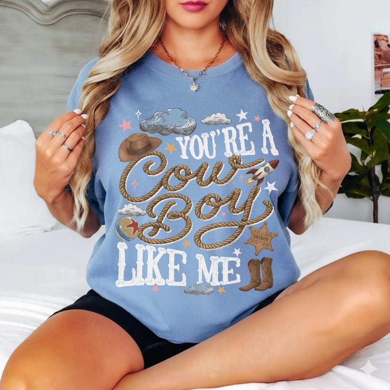 Puede incluir: Una camiseta azul claro de cuello redondo con el texto "YOU'RE A COWBOY LIKE ME" en letras blancas. El dise&ntilde;o incluye un sombrero de vaquero marr&oacute;n, una cuerda enrollada, un cohete peque&ntilde;o, una estrella de sheriff, una pelota y botas.