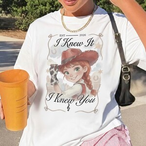 May include: A white t-shirt featuring a graphic of a cartoon cowgirl with red hair in braids and a brown hat. The text reads EST 2026, I Knew It, and I Knew You, framed by a rope design with horseshoe and star accents. A gold chain necklace is worn.