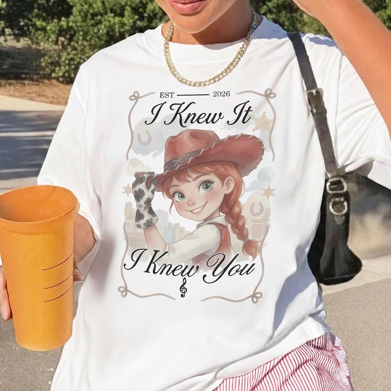 May include: A white t-shirt featuring a graphic of a cartoon cowgirl with red hair in braids and a brown hat. The text reads EST 2026, I Knew It, and I Knew You, framed by a rope design with horseshoe and star accents. A gold chain necklace is worn.
