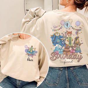 Pode incluir: Camisola bege com um gr&aacute;fico de desenho animado colorido. A frente apresenta um personagem de desenho animado azul com um chap&eacute;u de festa e o nome "Alex". A parte de tr&aacute;s tem um design "Birthday Squad" com personagens de desenhos animados, bal&otilde;es e o n&uacute;mero "4".